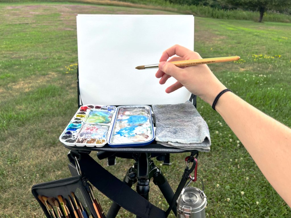 Image of a plein air easel set up with a blank watercolor block and open palette. A hand holding a brush prepares to make the first mark.