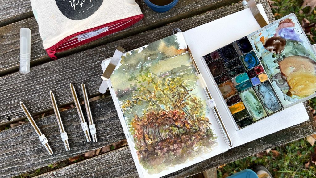 A watercolor sketch of some bittersweet branches in front of a pond. Autumn colors are the main focal point. A palette and assorted travel brushes are scattered around the sketchbook.